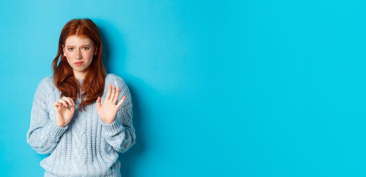 Reluctant redhead girl asking to stay away, shaking hand in rejection gesture, decline offer and grimacing displeased, standing over blue background.