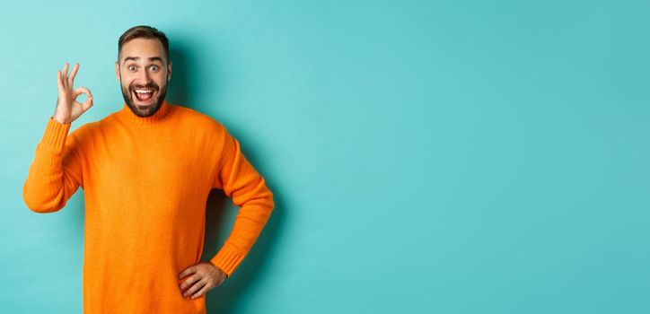 Impressed man recommending promo, showing okay sign and smiling amazed, approve and agree, standing over light blue background.