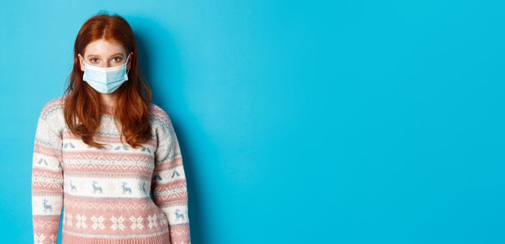 Winter, covid-19 and quarantine concept. Young redhead girl in sweater and face mask staring at camera, standing over blue background.