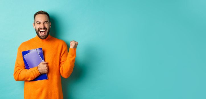 Education. Happy and satisfied man rejoicing, holding notebooks and making fist pump, shouting for joy, standing against turquoise background.