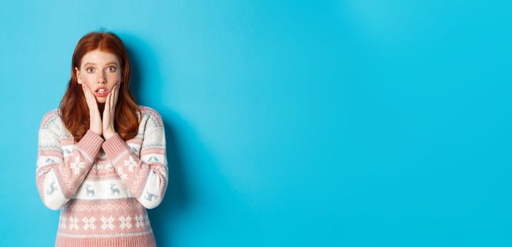 Image of impressed redhead girl gasping and staring at camera, hear big news, standing in winter sweater against blue background.