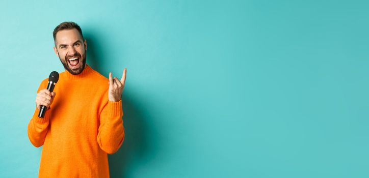 Joyful handsome man singing song in microphone, showing rock-n-roll gesture excited, standing over turquoise background.