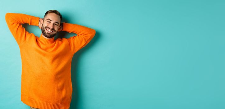 Handsome man relaxing, resting and smiling, holding hands behind head with carefree face, standing against light blue background.