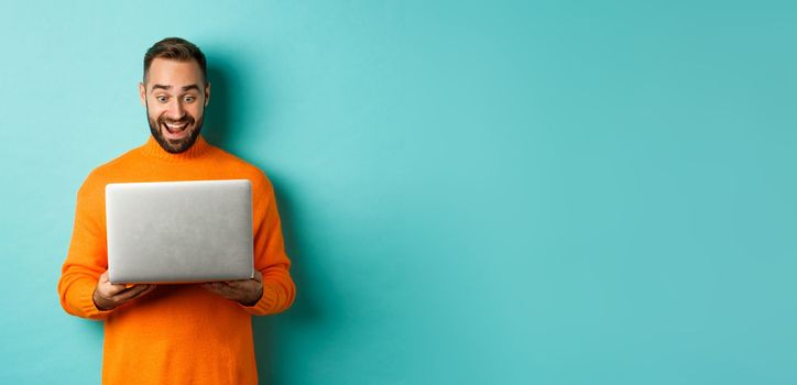 Excited man in orange sweater working on laptop, looking at computer screen amazed, standing over light blue background.