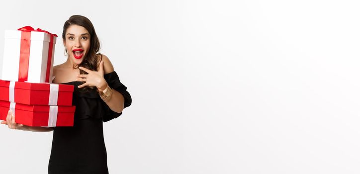 Celebration and christmas holidays concept. Excited and happy woman receive gifts, holding xmas presents and rejoicing, standing in black dress over white background.