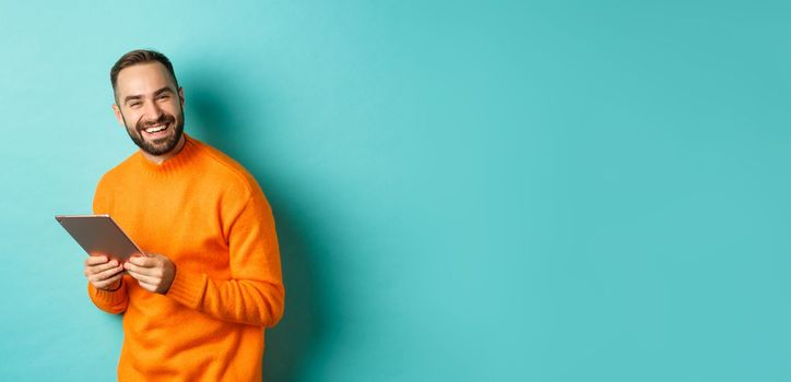 Handsome bearded man using digital tablet, laughing at camera, standing happy against light blue background.