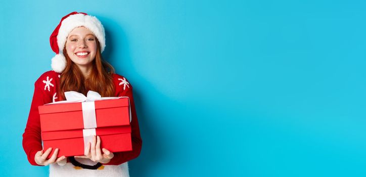 Happy holidays and Christmas concept. Cheerful teen girl with red hair, wearing santa hat and holding xmas presents.