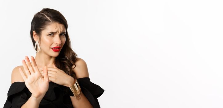 Fashion and beauty concept. Close-up of displeased young woman in black dress saying no, declining offer and looking with disdain, standing over white background.