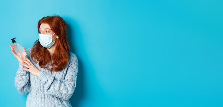 Winter, covid-19 and social distancing concept. Young redhead girl in face mask showing hand sanitizer, demonstrating antiseptic for disinfection, standing over blue background.