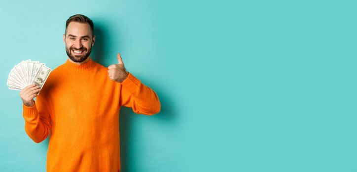 Shopping. Happy man showing thumb up and money in dollars, recommending bank loan or credits, standing over light blue background.