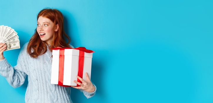 Christmas and shopping concept. Excited redhead girl looking at dollars, holding big New Year gift, buying presents, standing over blue background.