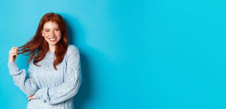 Flirty young woman with red hair, playing with hair and smiling, standing in sweater against blue background.