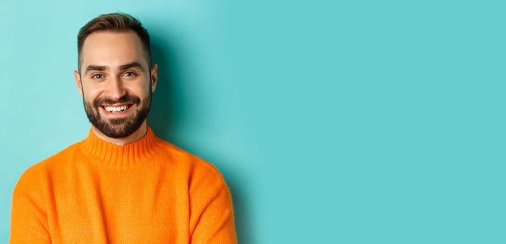 Close-up of handsome caucasian man smiling at camera, looking confident, wearing orange sweater, standing against turquoise background.
