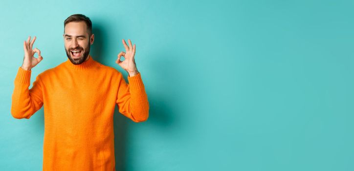 Confident man assuring all ok, showing okay signs and winking at you, recommending product, standing in orange sweater at studio background.