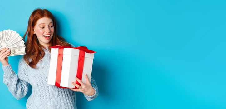 Christmas and shopping concept. Cheerful girl with red hair, holding money and big New Year gift, smiling happy, standing over blue background.