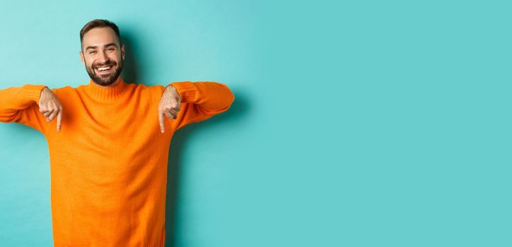 Happy adult man with beard pointing fingers down, smiling cheerful, showing advertisement, standing over turquoise background.