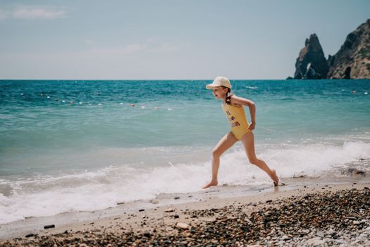 Cute little girl running along the seashore against a clear blue sea and rejoices in the rays of the summer sun. Beautiful girl in yellow swimsuit running and having fun on tropical beach