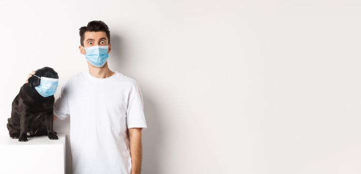 Covid-19, animals and quarantine concept. Image of funny young man and pug in medical masks, staring at camera, standing over white background.