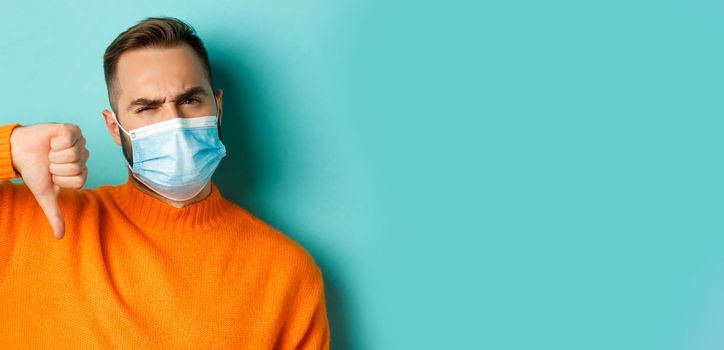 Covid-19, social distancing and quarantine concept. Close-up of disappointed male model in medical mask, showing thumb-down, negative feedback, standing over light blue background.