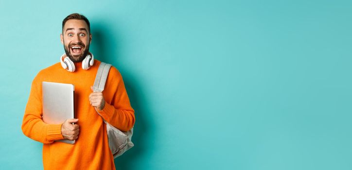 Happy man with backpack and headphones, holding laptop and smiling, looking excited, standing over turquoise background.