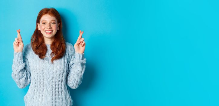 Hopeful redhead girl making a wish, cross fingers for good luck, smiling and anticipating good news or positive result, standing against blue background.