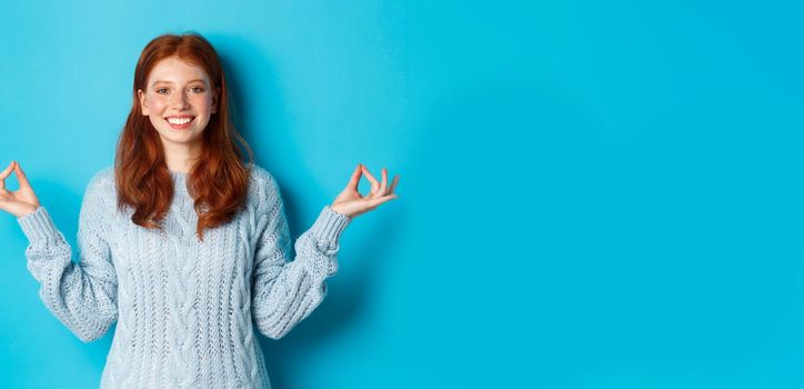 Smiling confident girl with red hair staying patient, holding hands in zen, meditation pose and staring at camera, practice yoga, standing calm against blue background.