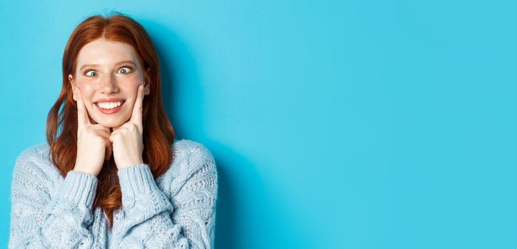 Close-up of funny redhead teen girl making faces, squinting and squeezing cheeks, standing against blue background.