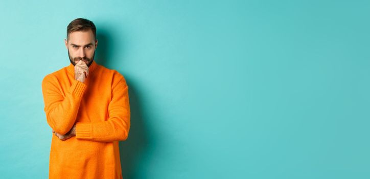 Thougtful young man making decision, looking serious and thinking, choosing, standing near copy space turquoise background.