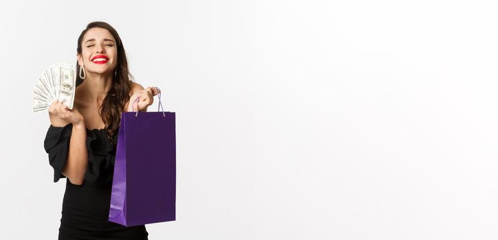 Satisfied and happy woman enjoying shopping, holding bag and money, smiling pleased, standing over white background.