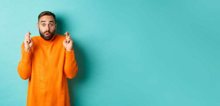 Hopeful man waiting with excitement, holding fingers crossed and making wish, standing over turquoise background.