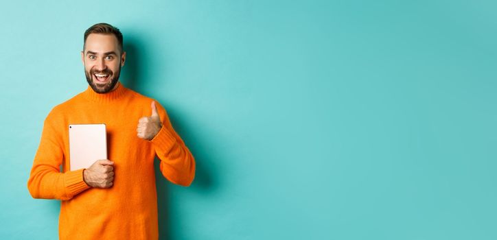 Work from home, technology concept. Handsome man holding laptop, showing thumb up, approve and like something, standing over turquoise background.