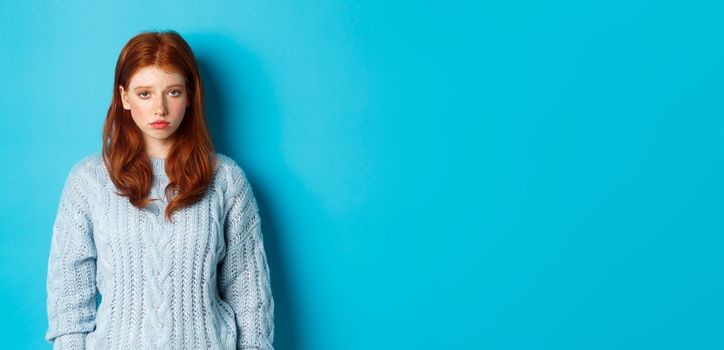 Sad and gloomy redhead teenage girl staring at camera uneasy, feeling bad, standing against blue backgorund in sweater.