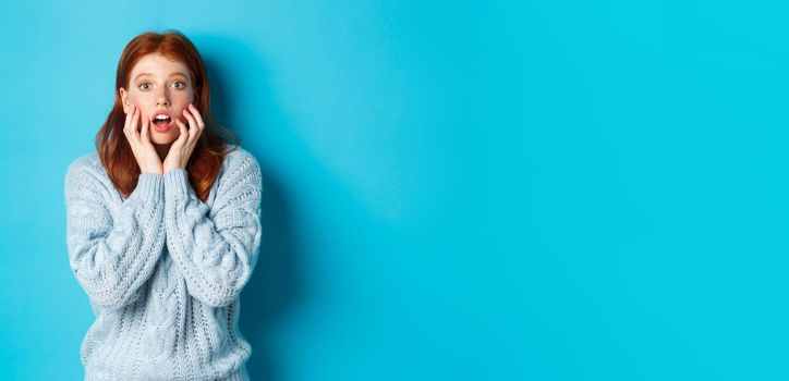 Startled redhead girl staring at something amazing, gasping in awe, standing in sweater against blue background. Copy space