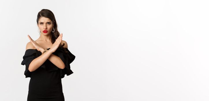 Fashion and beauty. Serious and confident female model in black dress, showing cross sign and frowning, stop gesture, telling no, standing over white background.