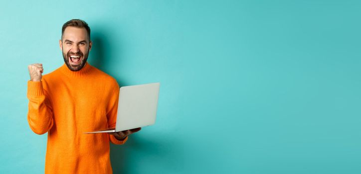 Freelance and technology concept. Lucky man winner celebrating, winning online, showing fist pump and holding laptop, standing over light blue background.