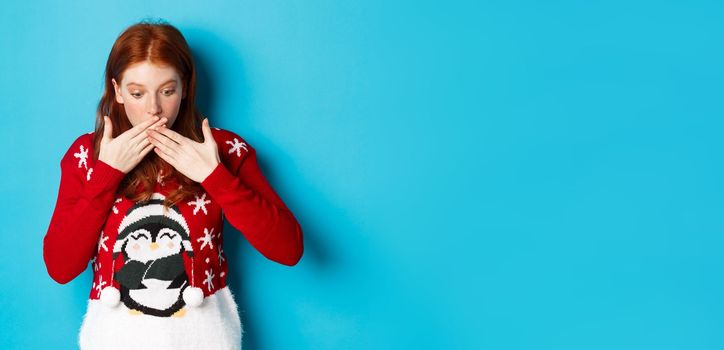 Winter holidays and Christmas Eve concept. Surprised redhead girl gasping, looking down with awe, staring at logo, standing in xmas sweater against blue background.