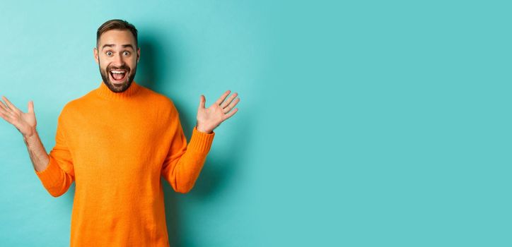 Excited man react to promo offer, spread hands sideways and rejoicing, standing against turquoise background in orange sweater.