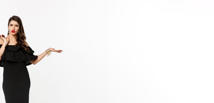 Beauty and fashion concept. Full length of arrogant and sassy woman in black dress, holding purse on shoulder and stroll forward at camera, posing over white background.