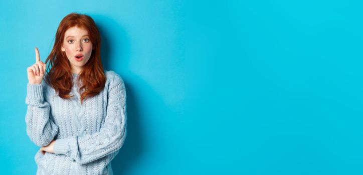 Thoughtful redhead girl having an idea, raising finger and saying suggestion, standing over blue background.