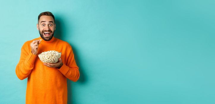 Excited young man watching interesting movie on tv screen, eating popcorn and looking amazed, blue background.