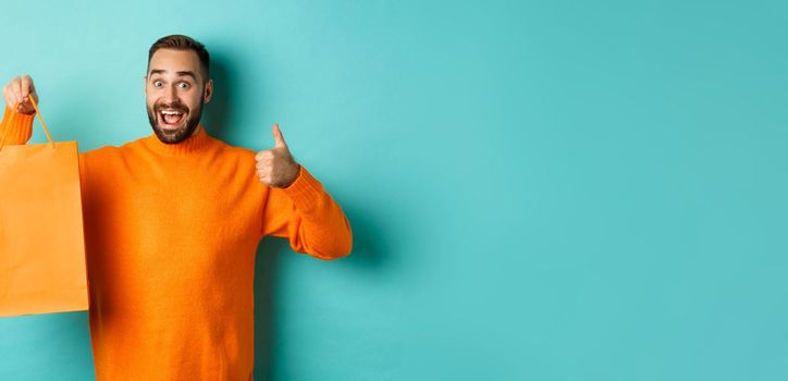 Happy man holding orange shopping bag and rejoicing from discount and celebrating, showing thumbs up and recommending, standing over blue background.