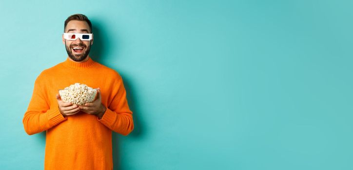 Happy guy watching movies in 3d glasses, eating popcorn and looking at camera, standing over light blue background. Copy space