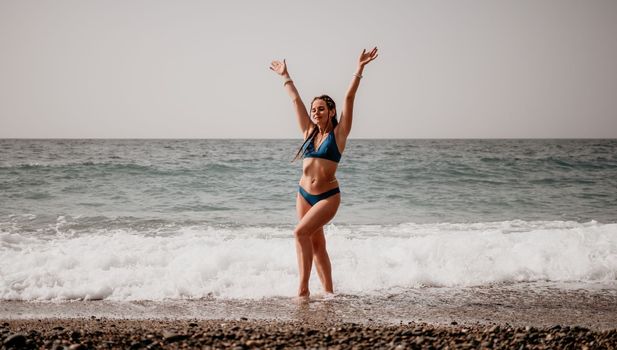 Beach vacation. Hot beautiful woman in sunhat and bikini standing with her arms raised to her head enjoying looking view of beach ocean on hot summer day.