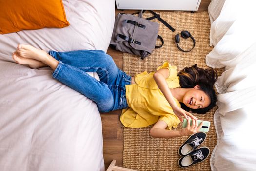 Top view of teen asian girl relaxing at home after class, lying on bedroom floor using phone. Adolescence, technology and lifestyle concepts.