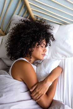 Top view of calm and serene young multiracial woman with curly hair sleeping in bed at home cozy bedroom. Vertical image. Lifestyle concept.