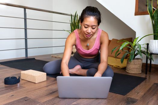 Young Asian woman using laptop to search online yoga class at home. Fitness and technology concept.