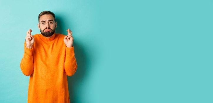 Hopeful sad man making a wish, cross fingers for good luck and sulking, standing over turquoise background.