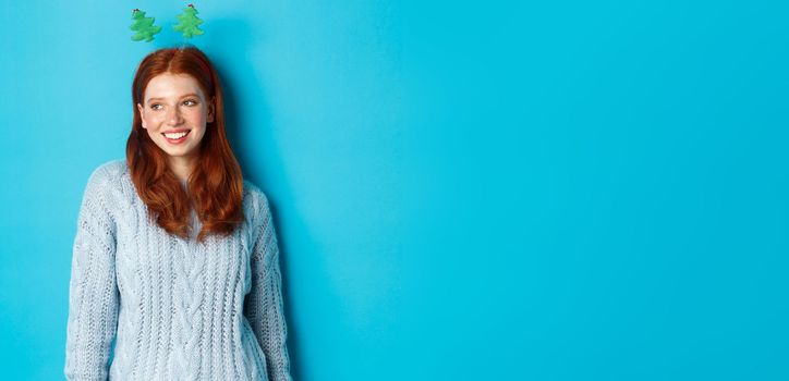 Winter holidays and Christmas sales concept. Cute redhead girl in funny New Year headband smiling, looking left at logo, standing over blue background.