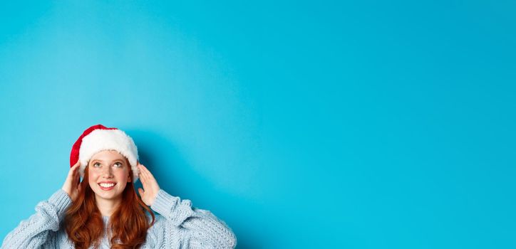 Winter holidays and Christmas eve concept. Head of cute redhead girl in santa hat, appear from bottom and looking up at copy space, staring logo, standing over blue background.