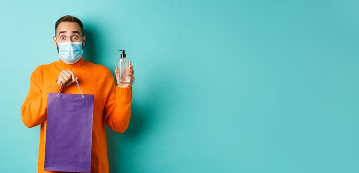 Coronavirus, pandemic and lifestyle concept. Man in face mask showing shopping bag and hand sanitizer, standing over turquoise background.